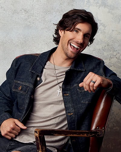 John King John King laughing and leaning against a leather chair on one arm wearing a jean jacket, gray t-shirt, and jeans.