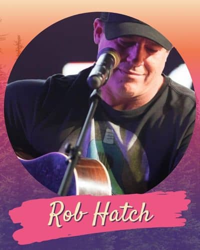 Rob Hatch playing a guitar wearing a black tee and baseball cap in front of a microphonr.